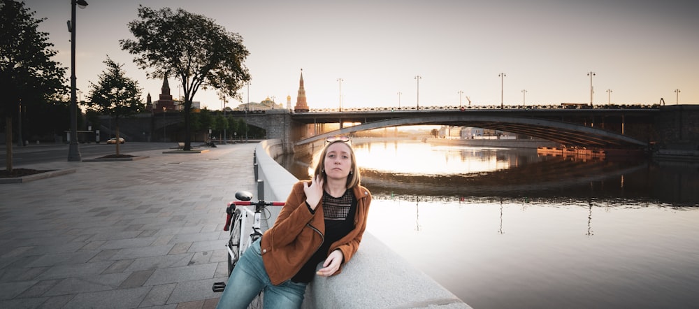 woman in brown leather jacket and blue denim jeans sitting on gray concrete bridge during daytime