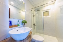 A modern bathroom featuring a glass-enclosed shower, a round ceramic sink on a stone countertop, and a mirror. There are toiletries in a woven basket, a liquid soap dispenser, and a small potted plant on the counter. The walls are finished with light-colored tiles, and there's a small window allowing natural light in.