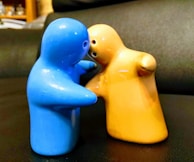 Two ceramic figurines resembling human shapes appear to be embracing or interacting closely. One figurine is blue, and the other is yellow-orange. They are positioned on a dark surface with a slightly blurred background, hinting at an indoor setting.