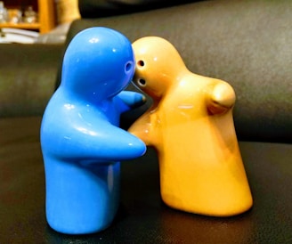 Two ceramic figurines resembling human shapes appear to be embracing or interacting closely. One figurine is blue, and the other is yellow-orange. They are positioned on a dark surface with a slightly blurred background, hinting at an indoor setting.