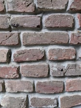 A close-up view of a brick wall, showcasing an arrangement of rectangular red-brown bricks with a rough texture. The bricks are laid in a horizontal pattern with visible mortar in between, displaying signs of weathering and small cracks.