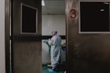 A person in medical attire stands in a room viewed through partially open double doors, numbered 3. The setting appears clinical, with a neutral color palette and a tiled ceiling. The person seems to be preparing or handling medical equipment or materials.