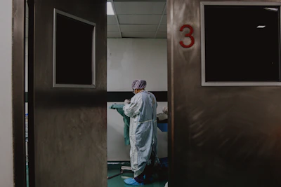 A person in medical attire stands in a room viewed through partially open double doors, numbered 3. The setting appears clinical, with a neutral color palette and a tiled ceiling. The person seems to be preparing or handling medical equipment or materials.