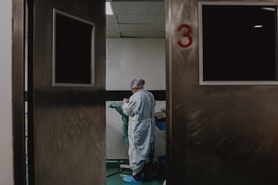 A person in medical attire stands in a room viewed through partially open double doors, numbered 3. The setting appears clinical, with a neutral color palette and a tiled ceiling. The person seems to be preparing or handling medical equipment or materials.