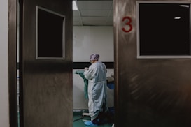 A person in medical attire stands in a room viewed through partially open double doors, numbered 3. The setting appears clinical, with a neutral color palette and a tiled ceiling. The person seems to be preparing or handling medical equipment or materials.