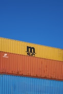Several stacked shipping containers with distinct colors, including yellow, orange, and blue, are visible against a clear blue sky. Industrial branding and logos are imprinted on the containers.
