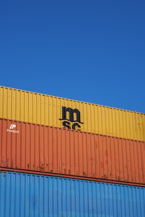 Several stacked shipping containers with distinct colors, including yellow, orange, and blue, are visible against a clear blue sky. Industrial branding and logos are imprinted on the containers.