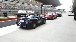 A row of luxury cars is parked alongside a modern racing track, with AMG branding visible on banners overhead. The setting appears to be a high-performance driving event, with the pit lane area and grandstands in the background.