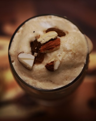 A frothy beverage topped with almond slices and raisins, presented in a transparent glass. The drink appears creamy with a smooth texture, and the almonds and raisins add a decorative and flavorful element.