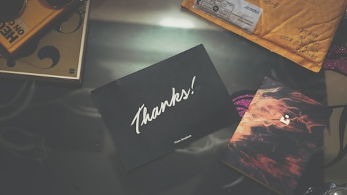 A black card with the word 'Thanks!' in white script is placed on a reflective surface. Surrounding it are various items including a brown envelope with handwritten information and a card with abstract designs featuring warm colors.