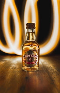 A bottle of Chivas Regal 12-year-old blended Scotch whisky is prominently placed on a wooden surface. Behind the bottle, there are bright, swirling light trails that create a dynamic and visually striking background. The bottle is filled with amber-colored liquid, and the label displays the brand name and product details.