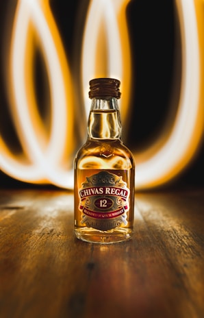 A bottle of Chivas Regal 12-year-old blended Scotch whisky is prominently placed on a wooden surface. Behind the bottle, there are bright, swirling light trails that create a dynamic and visually striking background. The bottle is filled with amber-colored liquid, and the label displays the brand name and product details.