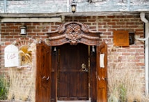 A rustic wooden door with intricate carvings is set in a red brick wall. The door has an old-fashioned knocker and is framed by an artistic wooden arch. To the left, there is a small window with decorative bars and a sign reading 'Cueva Materna'. A plaque on the wall indicates 'Caverna Materna'. Surrounding vegetation adds a natural and aged charm.