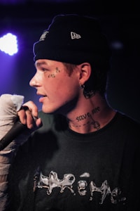 A person with tattoos on their face and neck holds a microphone, wearing a dark beanie and a black shirt with shiny lettering. They have earrings and a serious expression. The background is dimly lit with some purple light visible.