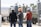 Three construction workers, wearing hard hats and casual work clothes, stand in front of industrial equipment and machinery outdoors. The background features large cylindrical tanks and various metal structures.