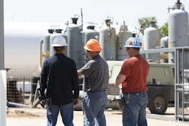 Three construction workers, wearing hard hats and casual work clothes, stand in front of industrial equipment and machinery outdoors. The background features large cylindrical tanks and various metal structures.