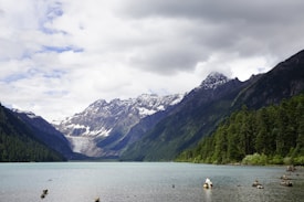 A tranquil mountainous landscape featuring a serene lake surrounded by dense evergreen forests. Snow-capped peaks tower in the background under a mostly cloudy sky, with patches of sunlight breaking through. A person in a kayak paddles near the lake's edge, adding a sense of scale and adventure to the scene.
