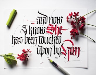 Elaborate calligraphy featuring a mix of black and red ink, with the text artfully arranged on a white paper. Surrounding the paper are small clusters of vibrant red flowers and a green leaf. A calligraphy pen with a green cap is placed beside the paper.