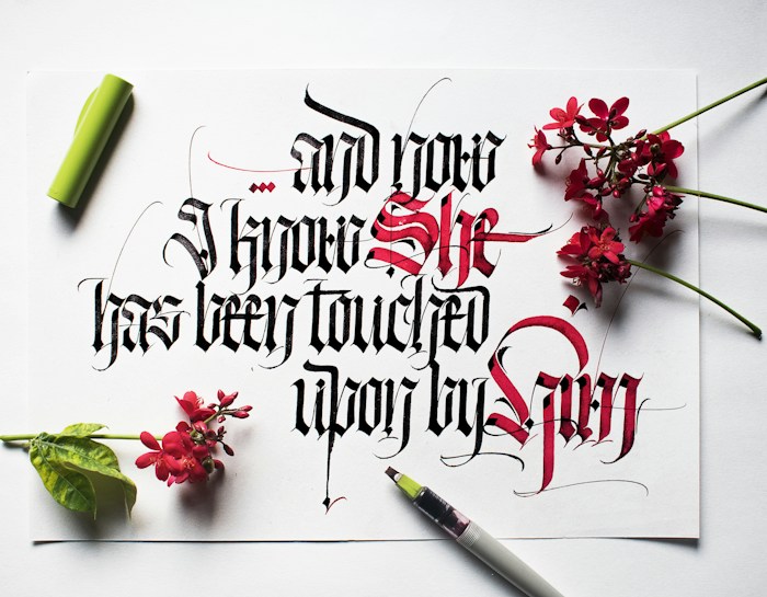 Elaborate calligraphy featuring a mix of black and red ink, with the text artfully arranged on a white paper. Surrounding the paper are small clusters of vibrant red flowers and a green leaf. A calligraphy pen with a green cap is placed beside the paper.