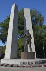 A monument featuring a statue of a soldier in uniform, equipped with a gun, set against two tall, concrete pillars. The words 'СЛАВА ГЕРОЯМ' are inscribed on the base. The background consists of green trees under a clear blue sky.