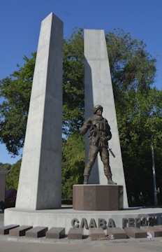 A monument featuring a statue of a soldier in uniform, equipped with a gun, set against two tall, concrete pillars. The words 'СЛАВА ГЕРОЯМ' are inscribed on the base. The background consists of green trees under a clear blue sky.