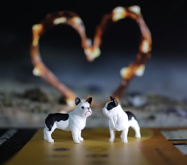 Two small figurines of black and white bulldogs are placed prominently in the foreground. Behind them, there is a soft-focus background featuring a heart shape formed by warm, glowing string lights. The scene has a bokeh effect, with the background lights appearing blurred and creating an atmospheric depth.