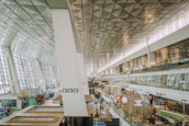 A spacious and modern airport terminal with large glass windows and a high ceiling. Various signs and advertisements for shops and food outlets can be seen. The area is well-lit with natural and artificial light, and there are people walking and standing in different parts of the terminal.
