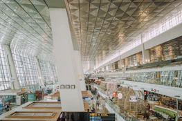 A spacious and modern airport terminal with large glass windows and a high ceiling. Various signs and advertisements for shops and food outlets can be seen. The area is well-lit with natural and artificial light, and there are people walking and standing in different parts of the terminal.