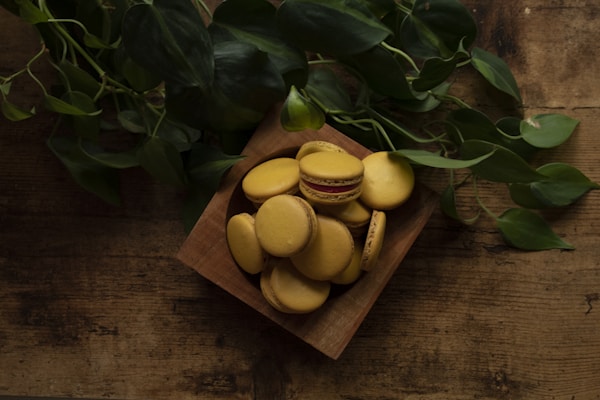 A wooden bowl filled with yellow macarons, placed on a rustic wooden surface. Lush green leaves partially surround the bowl, adding a natural element to the setting. The lighting is soft and creates a cozy, earthy ambiance.