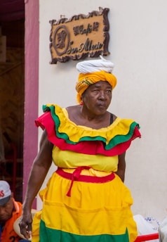 An individual in vibrant traditional clothing comprising yellow, red, and green hues stands in front of a cafe sign mounted on a beige wall. The outfit is complemented with a colorful headwrap and appears to be a cultural or festive attire.
