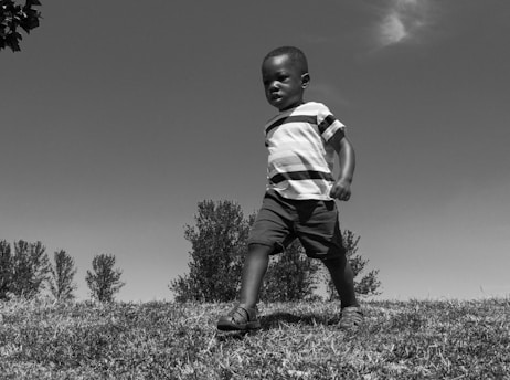 A young child strides confidently across a grassy field. The black-and-white image highlights the contrast between the child's striped shirt and the clear sky above. Trees in the background create a natural, serene setting.