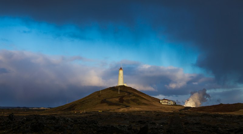 A white lighthouse on a brown hill under dark storm clouds, with geothermal steam billowing from the ground to its right