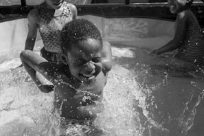 Children are playing joyfully in a small inflatable pool, splashing water around. The boy in the center is squinting and smiling as water splashes around him. Two other children are partially visible, also enjoying the water play.