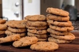 Several stacks of homemade chocolate chip cookies are neatly piled on a wooden surface. Each cookie has a golden-brown color and is speckled with visible chocolate chips. The background shows a slightly blurred kitchen area, hinting at a domestic setting.