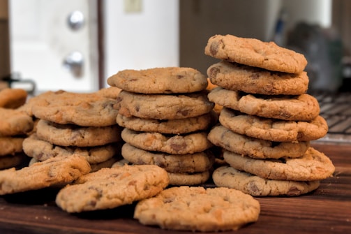 Several stacks of homemade chocolate chip cookies are neatly piled on a wooden surface. Each cookie has a golden-brown color and is speckled with visible chocolate chips. The background shows a slightly blurred kitchen area, hinting at a domestic setting.