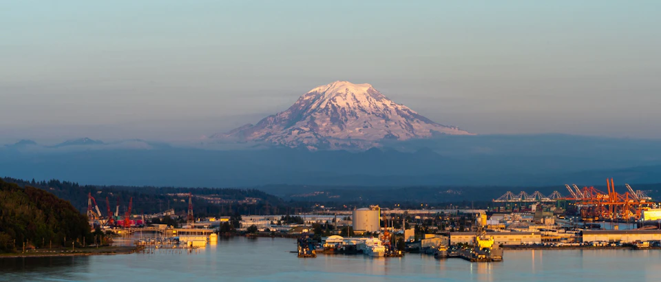 The image depicts a serene harbor scene with a large mountain range in the background, its snow-capped peak contrasting against the clear blue sky. The calm waters of the harbor are dotted with several boats and cranes, suggesting ongoing construction or industrial activity nearby. The perspective is from above, providing a comprehensive view of the cityscape nestled at the foot of the mountains.