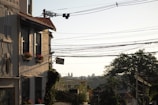 A residential street scene with a house on the left featuring flower boxes filled with red and pink flowers. Numerous overhead electric wires stretch across the sky. In the background, distant buildings are visible among a hazy skyline, with trees lining the streets.