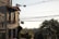 A residential street scene with a house on the left featuring flower boxes filled with red and pink flowers. Numerous overhead electric wires stretch across the sky. In the background, distant buildings are visible among a hazy skyline, with trees lining the streets.