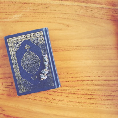A beautifully ornate book with intricate gold embellishments lies closed on a light wooden surface. Sprigs of dried lavender are placed gracefully on top of the book, adding a touch of nature and simplicity.