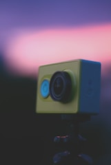A close-up view of a yellow action camera mounted on a tripod with an out-of-focus background featuring a gradient of purple and pink hues.