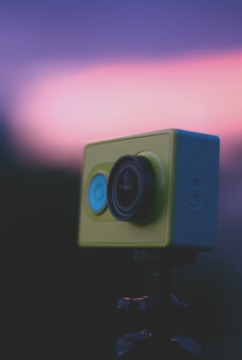A close-up view of a yellow action camera mounted on a tripod with an out-of-focus background featuring a gradient of purple and pink hues.