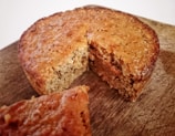 A golden-brown baked cake with a textured crust sits on a wooden surface. A slice has been cut and slightly separated from the main cake, revealing a moist and soft interior.