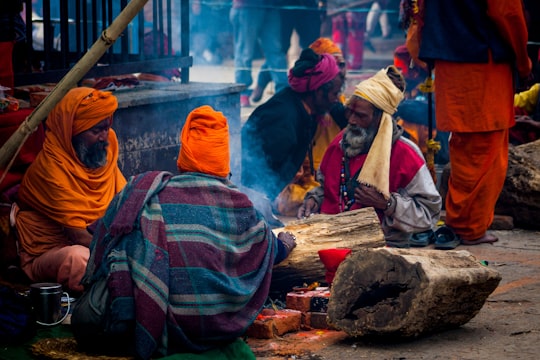 people in orange and white hijab sitting on brown wooden log during daytime in Pashupatinath Nepal