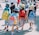 A group of young children wearing colorful backpacks and hats walk together in a line, possibly on a school outing. They are accompanied by an adult. The scene is lively, with bright sunlight casting shadows on the ground.