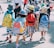 A group of young children wearing colorful backpacks and hats walk together in a line, possibly on a school outing. They are accompanied by an adult. The scene is lively, with bright sunlight casting shadows on the ground.