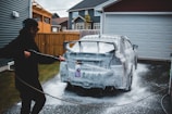 A person is washing a car with a pressure washer, applying soap foam all over the vehicle. The scene takes place in a driveway next to a garage, with suburban houses in the background. The person is wearing a cap and appears to be focused on cleaning the car.