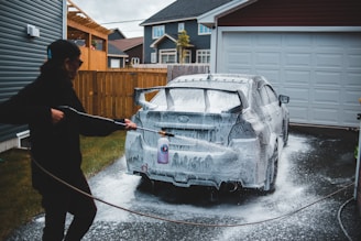 A person is washing a car with a pressure washer, applying soap foam all over the vehicle. The scene takes place in a driveway next to a garage, with suburban houses in the background. The person is wearing a cap and appears to be focused on cleaning the car.
