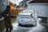 A person is washing a car with a pressure washer, applying soap foam all over the vehicle. The scene takes place in a driveway next to a garage, with suburban houses in the background. The person is wearing a cap and appears to be focused on cleaning the car.