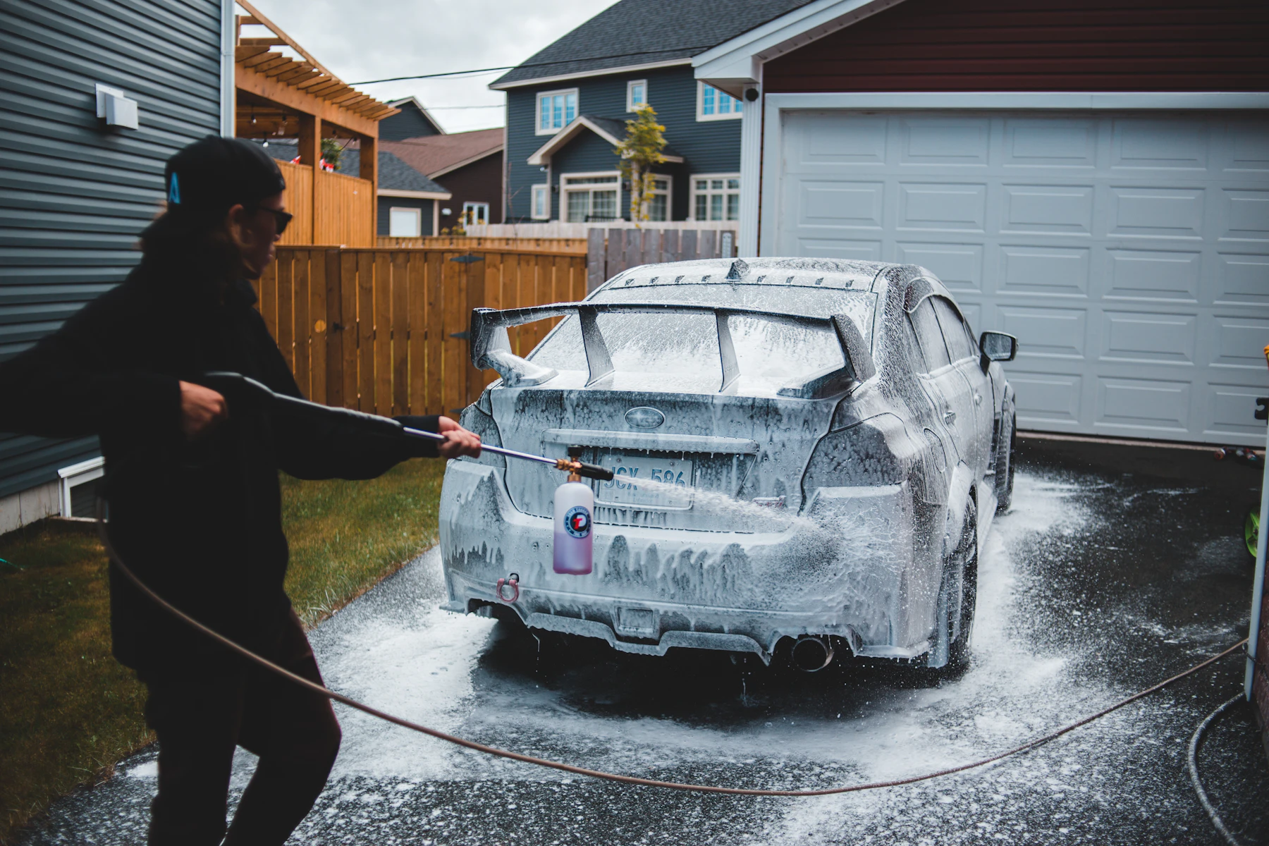 A person rinsing a foam-covered silver car on a driveway beside a garage