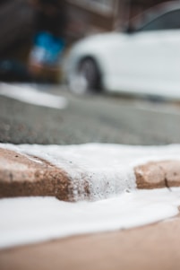 A close-up view of white foam on a concrete surface, with a blurred background featuring part of a white car. The focus is on the texture and flow of the foam.